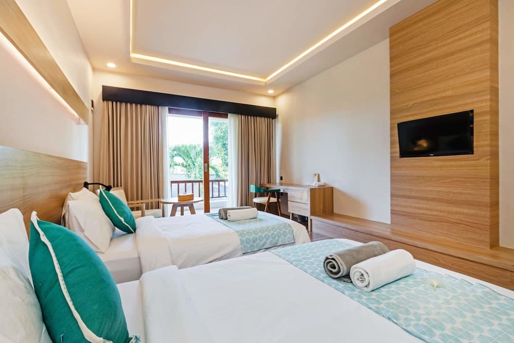 A cozy hotel room with two neatly made beds adorned with turquoise accents, a wooden desk, and a balcony offering lush greenery views, ideal for travelers seeking comfort and relaxation.