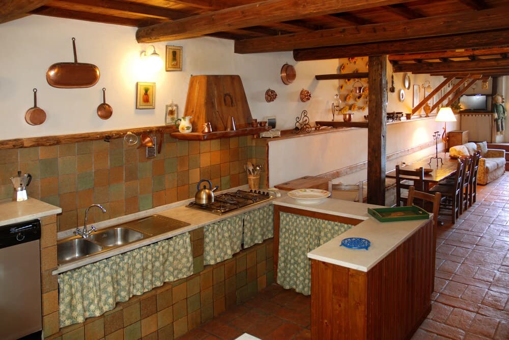 A cozy rustic kitchen with wooden beams, tiled walls, hanging copper pots, and a warm ambiance invites travelers to experience traditional culinary settings in a charming countryside retreat.