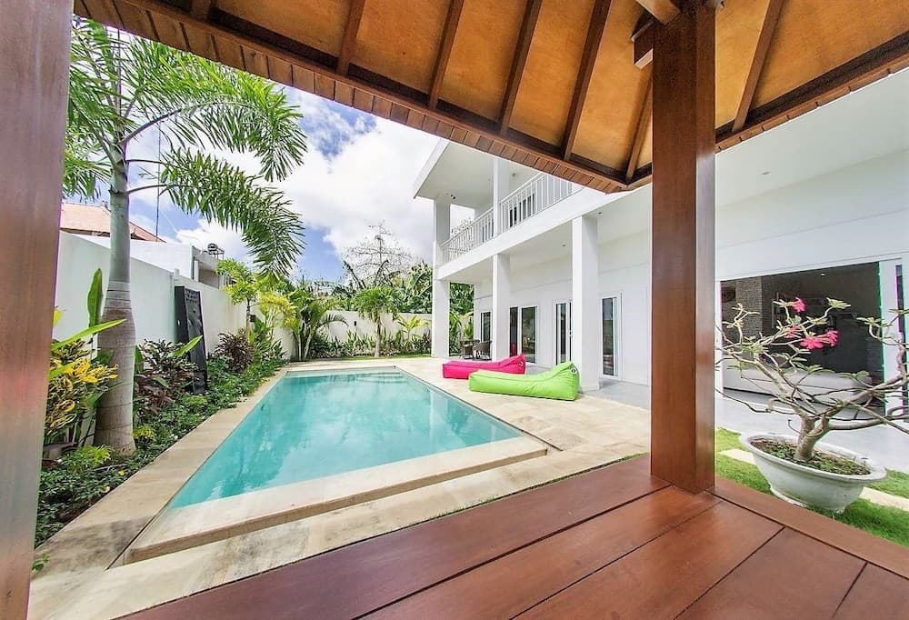 A serene tropical villa courtyard features a tranquil pool, vibrant bean bags, lush greenery, and a wooden pavilion, ideal for a relaxing vacation retreat.
