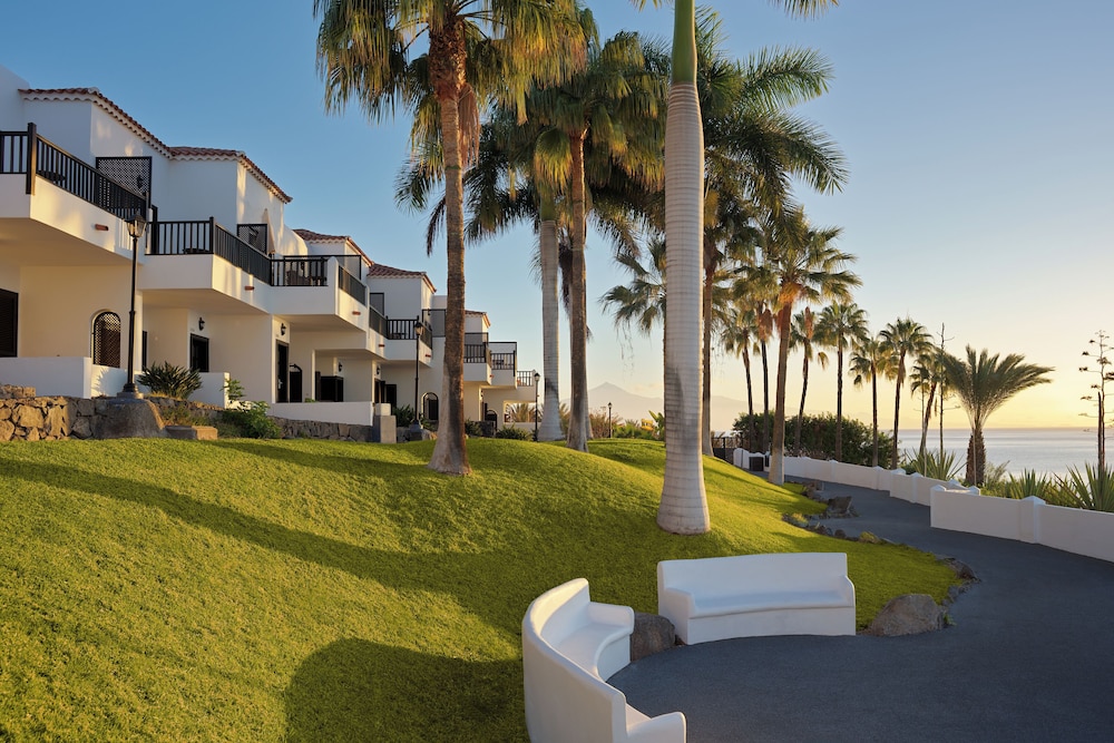 A serene coastal resort features white villas with balconies overlooking lush green lawns lined with tall palm trees, creating an inviting atmosphere perfect for relaxation and leisure travel.