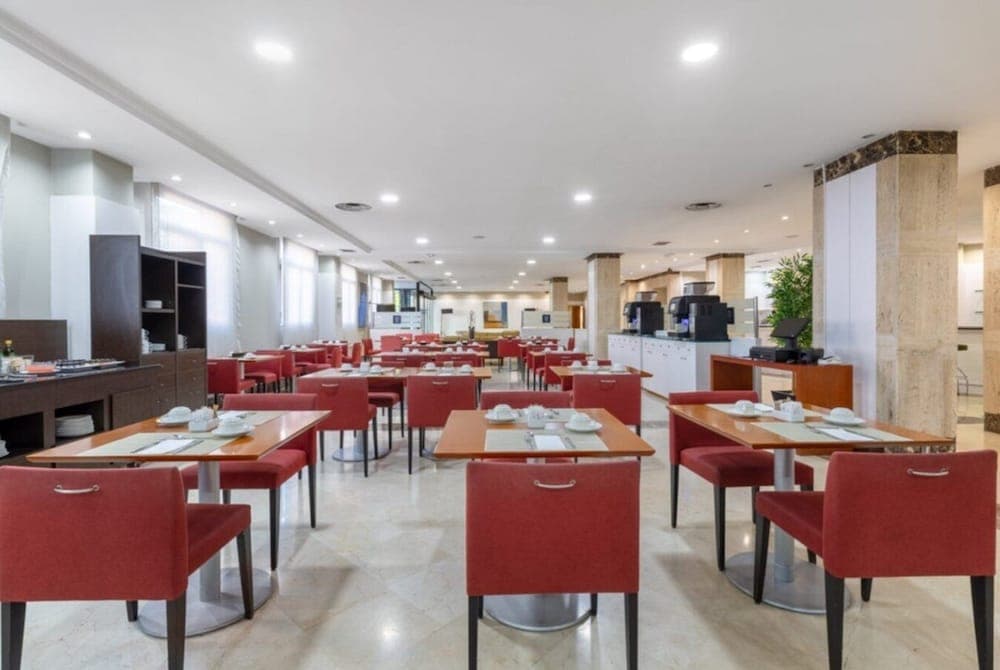 A spacious hotel dining area with red chairs, wooden tables set for breakfast, bright lighting, and modern decor offers travelers a welcoming and functional space for morning meals.