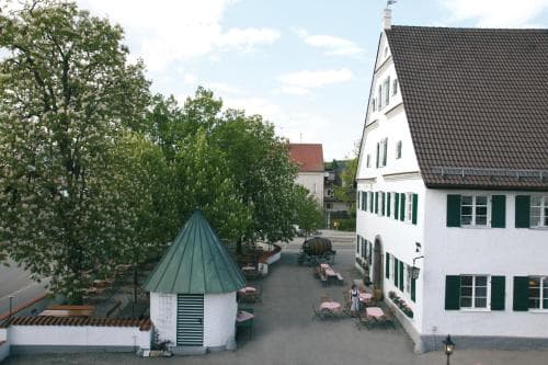 A quaint European village square features a charming white building with green shutters, outdoor seating, and a small gazebo surrounded by lush trees, ideal for a peaceful getaway or cultural exploration.