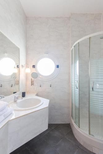 A serene and modern bathroom features a sleek glass shower enclosure, a minimalist vanity with neatly folded towels, and a round window that bathes the space in soft natural light, ideal for travelers seeking a luxurious and relaxing retreat.