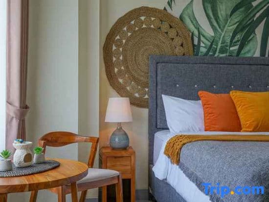 A cozy hotel room features a neatly made bed adorned with orange pillows, a woven wall decoration, and a wooden table set by the window, ideal for travelers seeking comfort and relaxation.