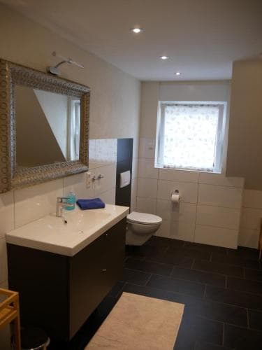 A modern bathroom with sleek black tiles, a white vanity sink, a toilet, and a frosted window creates a clean and serene atmosphere, ideal for travelers seeking comfort and style in their accommodations.