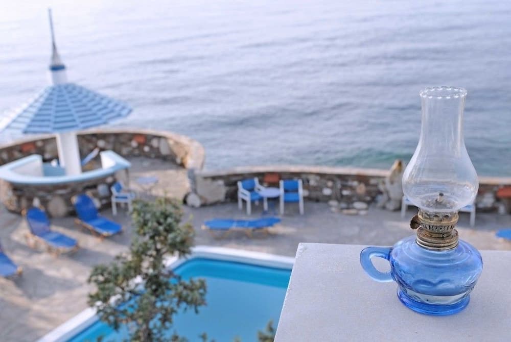 A serene seaside retreat features a tranquil poolside area with blue lounge chairs, a stone wall overlooking the ocean, and a vintage blue oil lamp in the foreground, perfect for a relaxing vacation escape.