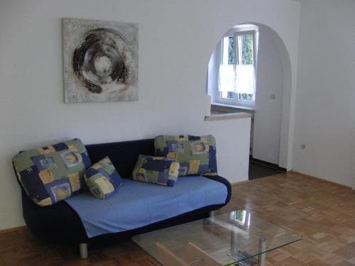 A cozy living room with a blue sofa adorned with patterned cushions, a glass coffee table, and an arched window letting in natural light, creates a serene atmosphere ideal for a relaxing vacation rental stay.