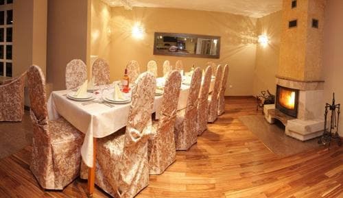 A cozy dining room with a long table set for a gathering, surrounded by upholstered chairs, features warm lighting, a fireplace, and wooden floors, ideal for intimate family dinners or small celebrations.