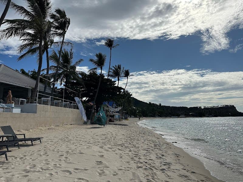 A serene tropical beach scene at Tembo Beach Club & Resort in Thailand, featuring sandy shores, palm trees, lounge chairs, and a calm sea under a partly cloudy sky, ideal for relaxation and leisure travel.