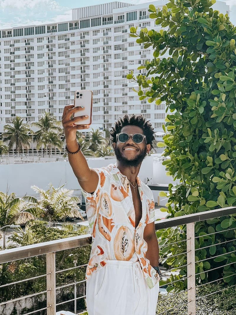 A joyful traveler captures a selfie on a sunny balcony overlooking lush greenery and towering hotel buildings in Miami Beach, perfect for showcasing vibrant vacation vibes.