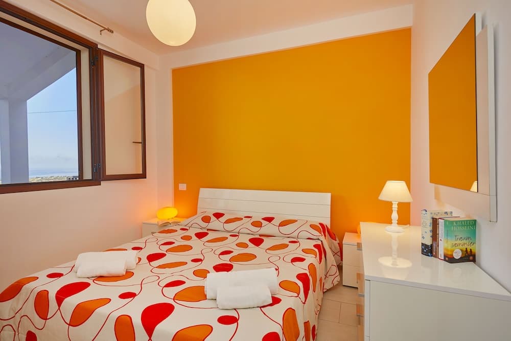 A cozy bedroom with an orange accent wall, patterned bedding, and a scenic view through the window offers a relaxing retreat for travelers seeking comfort and style.