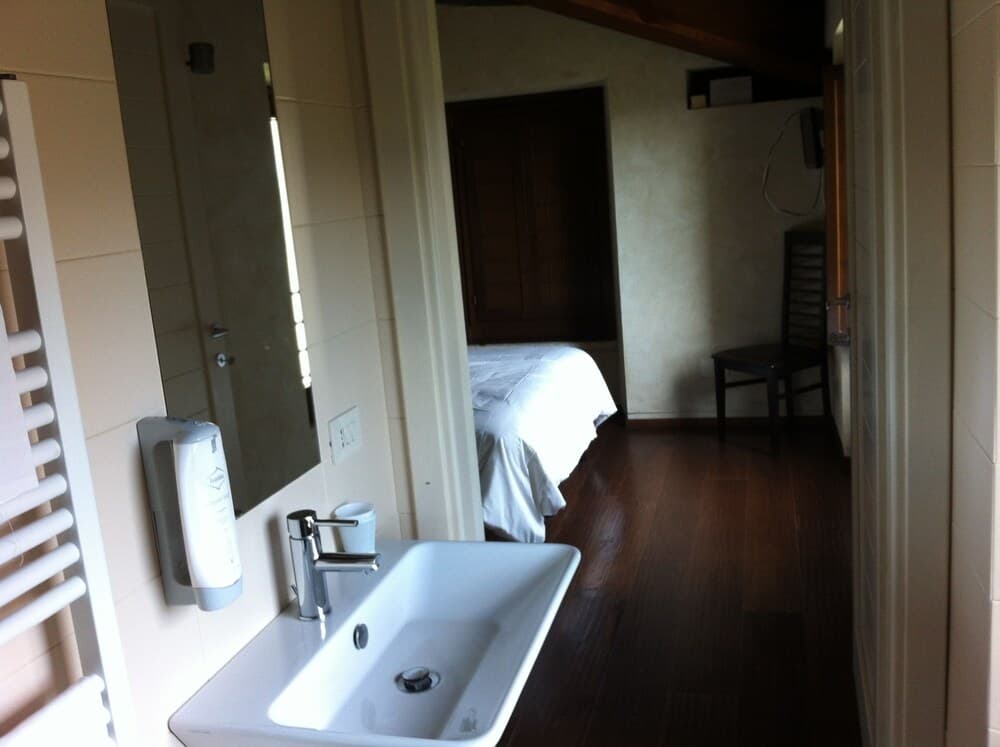 A cozy hotel room with a bathroom sink, mirror, towel radiator, and a view into the bedroom featuring wooden floors, a bed, and chair, ideal for travelers seeking comfort and simplicity.
