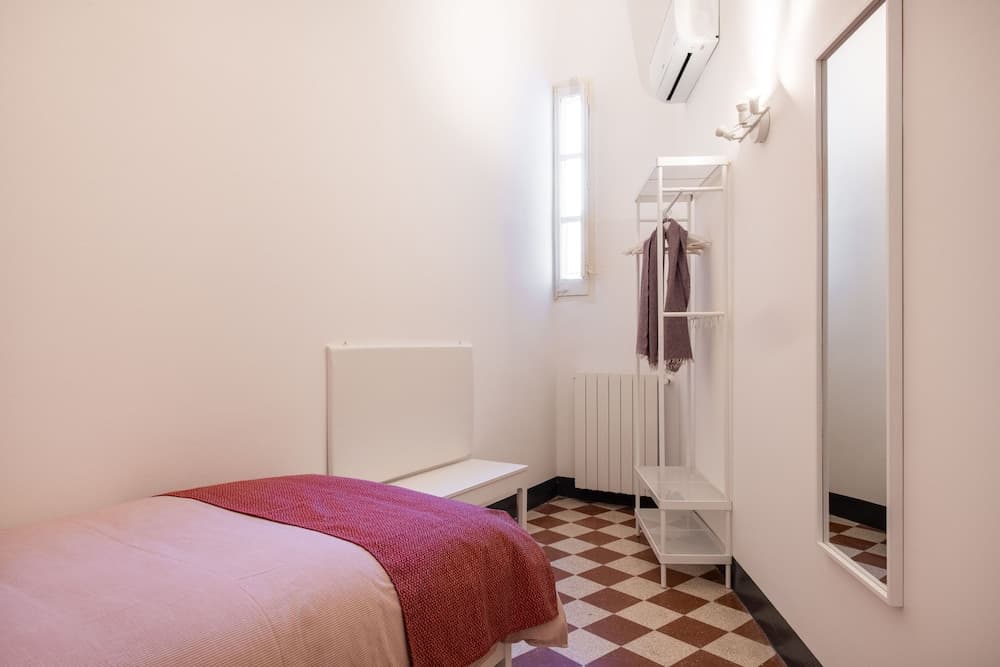 A cozy minimalist bedroom with a neatly made bed, checkered floor tiles, a white shelving unit, and a full-length mirror, ideal for travelers seeking a clean and simple accommodation option.