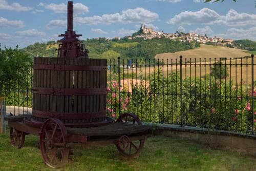 A rustic wooden wine press stands in the foreground of a picturesque hillside landscape, overlooking a charming village perched atop a green hill under a partly cloudy sky, ideal for travelers seeking cultural heritage and scenic beauty.