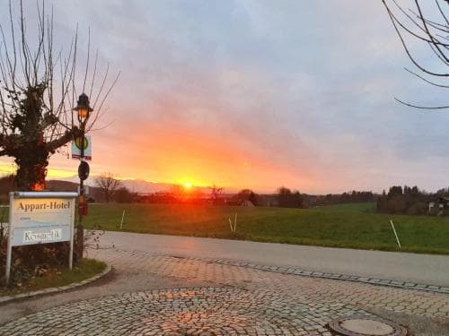 A serene sunset casts warm hues over a cobblestone driveway leading to an Appart-Hotel sign, surrounded by tranquil fields, ideal for travelers seeking a peaceful countryside retreat.