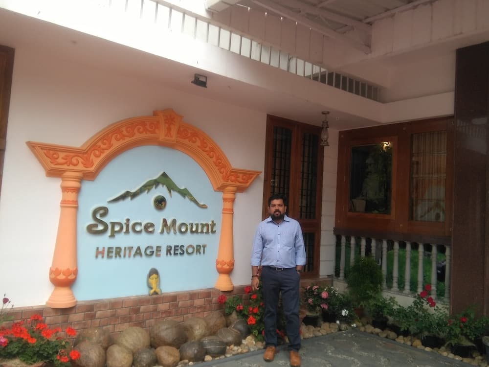 A traveler stands proudly in front of the vibrant entrance sign of Spice Mount Heritage Resort, surrounded by lush greenery and colorful flowers, ideal for a serene getaway experience.