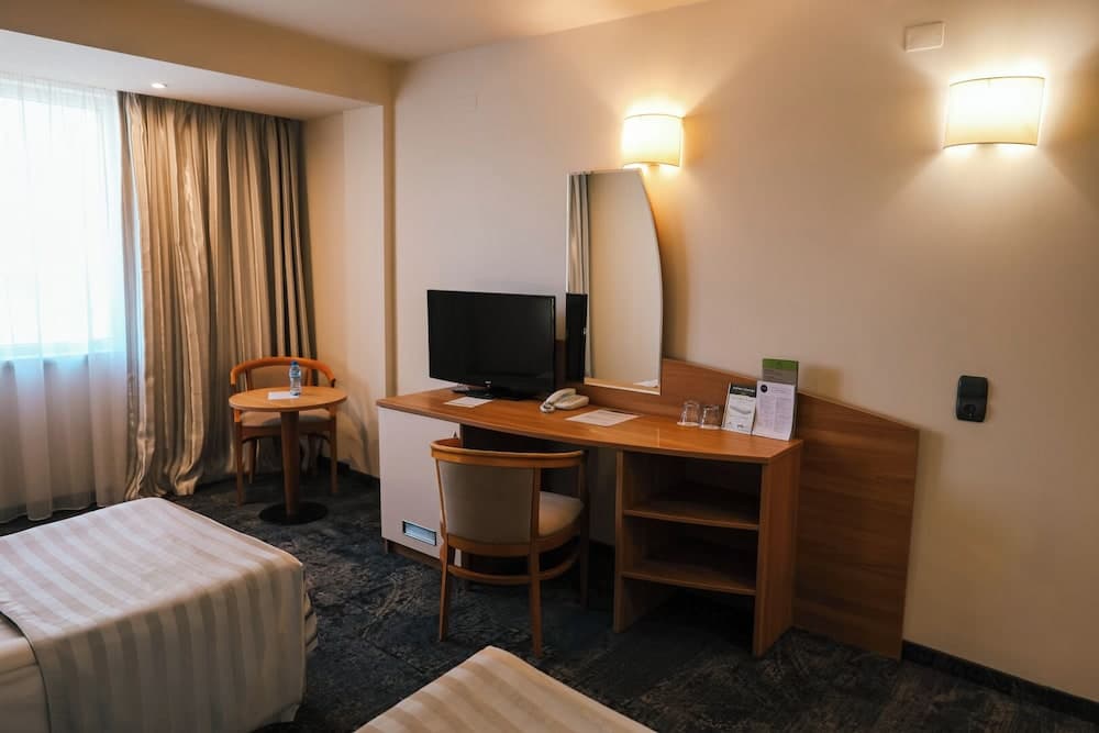 A cozy hotel room features a neatly arranged desk with a TV, mirror, and comfortable seating by a window with curtains, ideal for travelers seeking a functional and relaxing stay.