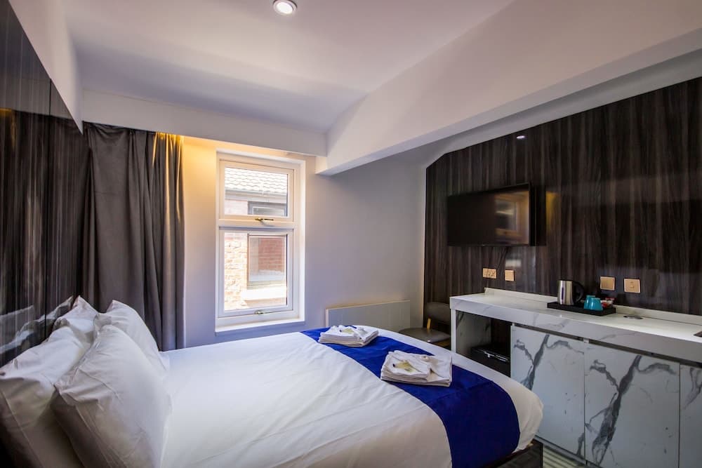 A cozy hotel room features a neatly made bed by a window with brick building views, a sleek marble counter with tea setup, and a modern TV, ideal for travelers seeking comfort and convenience.