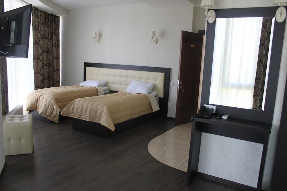 A cozy hotel room features two neatly arranged twin beds, a desk with a phone, soft lighting from wall sconces, and large windows with patterned curtains, ideal for travelers seeking comfort and functionality.