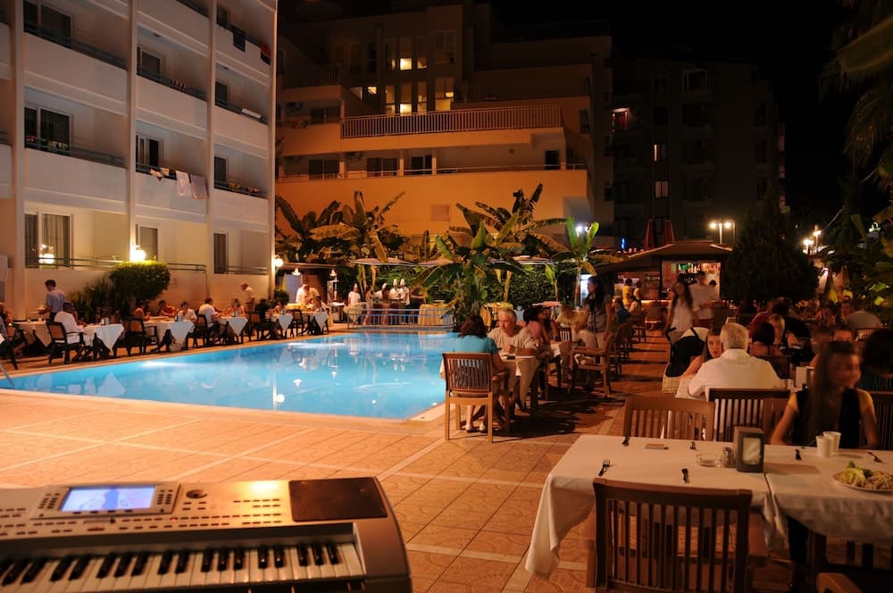 A cozy evening at an outdoor dining area by a poolside, illuminated by warm lights, surrounded by lush greenery and apartment buildings, featuring tables set for dinner and a keyboard ready for live music, perfect for a relaxing vacation getaway.