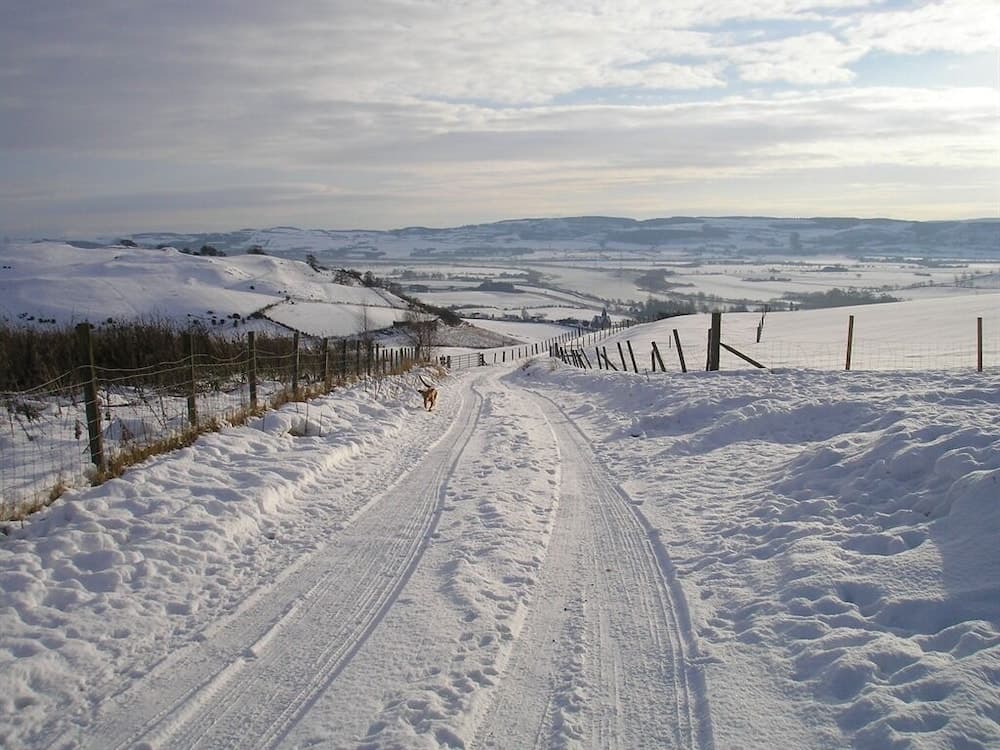 A serene winter landscape features a snow-covered rural road flanked by fences, with distant rolling hills under a partly cloudy sky, ideal for winter hiking or scenic drives.