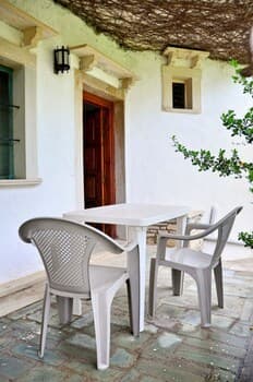 A serene outdoor patio features a simple white table and chairs under a rustic thatched roof, ideal for travelers seeking a peaceful retreat in a quaint countryside setting.