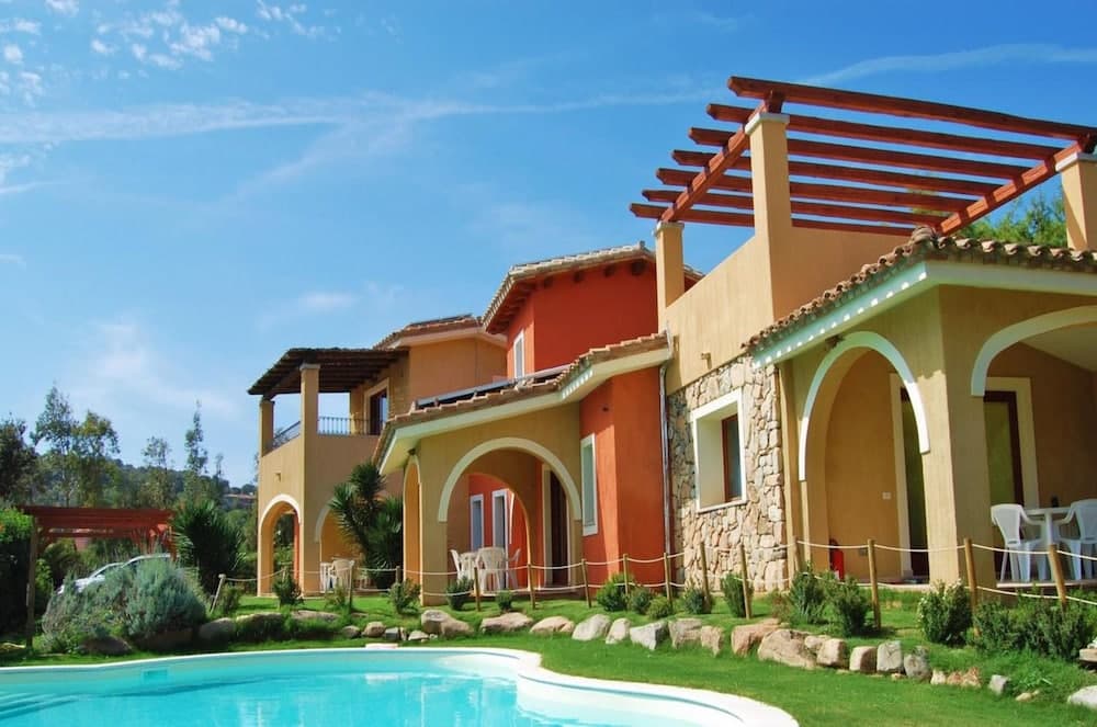 A vibrant Mediterranean villa with arched doorways, a wooden pergola, and a serene poolside garden under a clear blue sky, ideal for a relaxing family vacation.