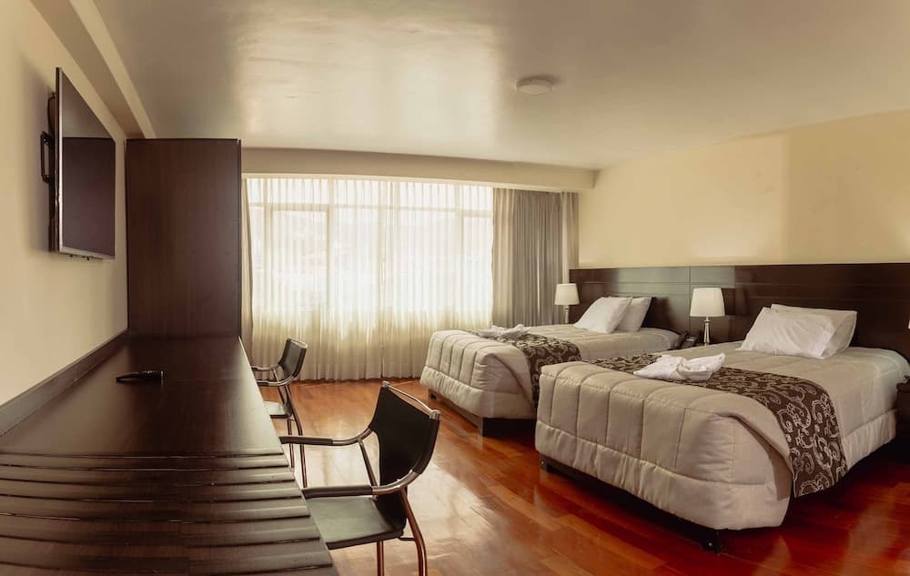 A cozy hotel room with two neatly made beds, a wooden desk, chairs, and a large window with sheer curtains, ideal for travelers seeking comfort and functionality.
