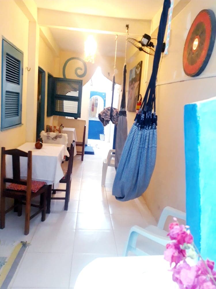 A cozy indoor dining area with white tiled floors, wooden tables draped in white cloths, hanging blue hammocks, and vibrant wall decorations creates a relaxed and inviting atmosphere perfect for travelers seeking a unique local experience.