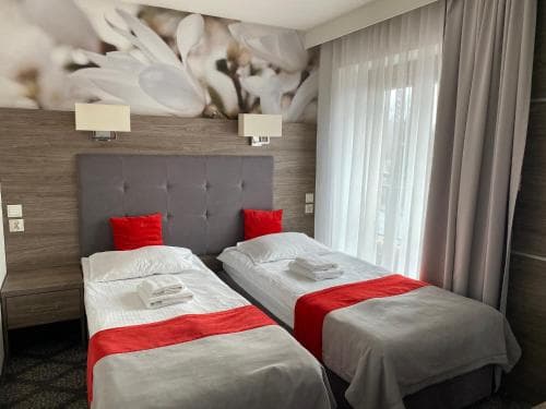 A cozy hotel room features two neatly made twin beds with red accents against a floral wall mural, creating a welcoming atmosphere ideal for travelers seeking comfort and relaxation.