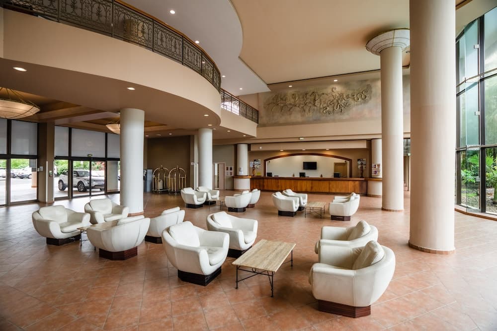 A spacious hotel lobby with elegant white armchairs, tiled floors, and large windows exudes a welcoming atmosphere, ideal for travelers seeking comfort and relaxation during their stay.