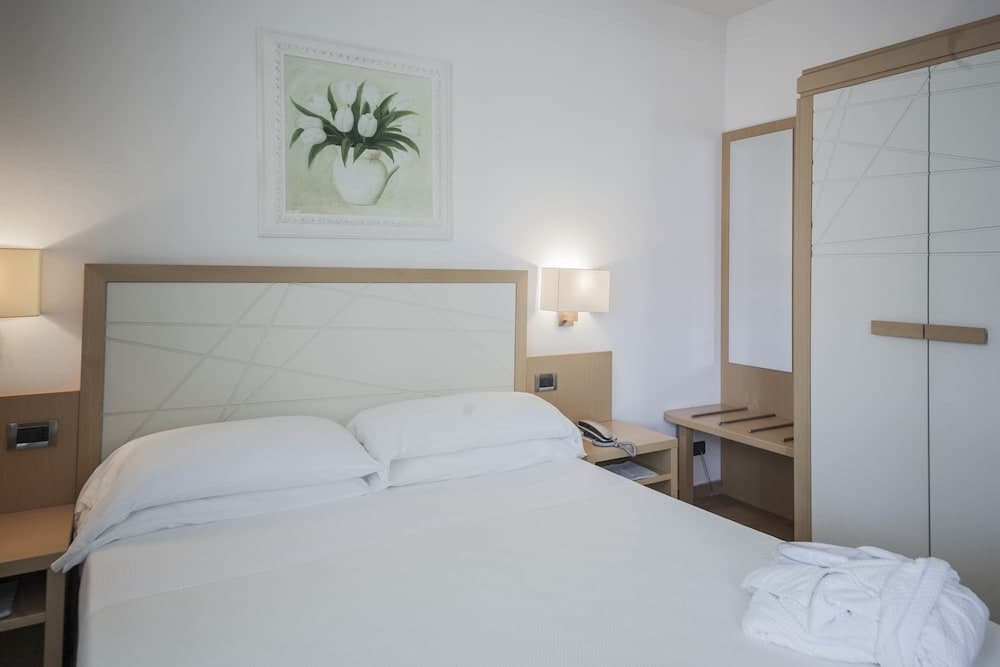 A cozy hotel room features a neatly made bed with white linens, wooden bedside tables with lamps, a floral painting above the bed, and a small desk with a mirror, ideal for travelers seeking comfort and simplicity.
