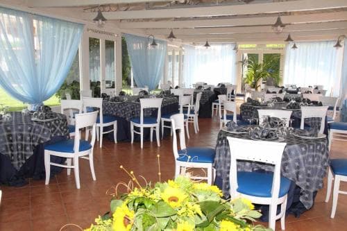A spacious indoor dining area with blue-curtained windows, white chairs, patterned tablecloths, and vibrant sunflowers creates an inviting atmosphere ideal for wedding receptions or group gatherings.