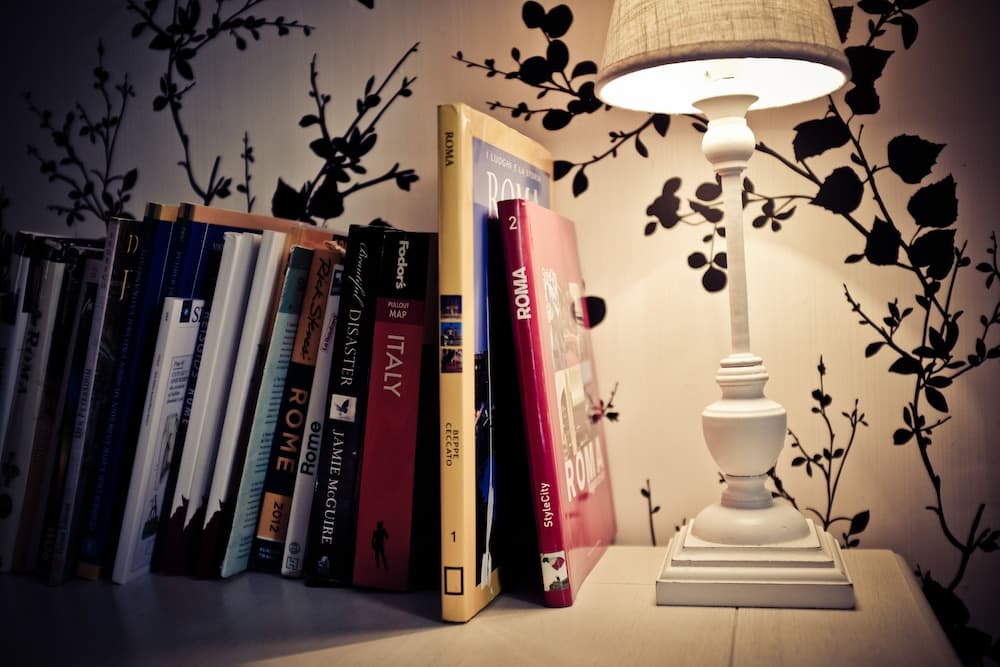A cozy reading nook features travel guides on Rome and Italy neatly arranged beside a glowing white lamp against a leaf-patterned wall, ideal for travelers planning their next adventure.