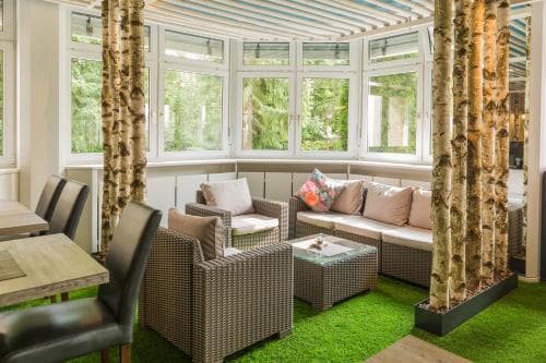 A cozy indoor lounge area with artificial turf flooring, birch tree pillars, and large windows offering a serene forest view, ideal for relaxation or a tranquil retreat destination.
