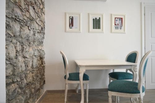 A cozy dining nook features a white table with teal-cushioned chairs against a rustic stone wall, framed by minimalist artwork, ideal for travelers seeking charming accommodations with a blend of modern comfort and traditional charm.