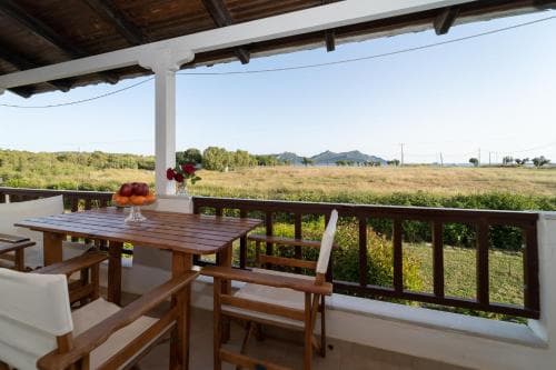 A serene balcony overlooks a lush countryside with distant hills, featuring a cozy dining setup and vibrant flowers, ideal for a tranquil rural retreat.