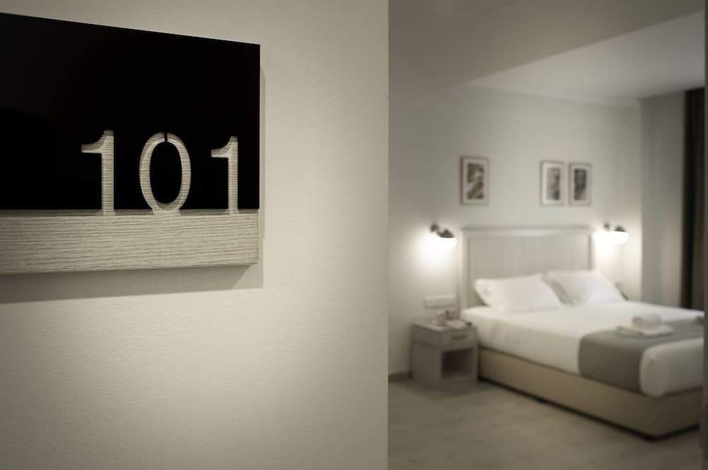 A serene hotel room with a minimalist design features a neatly made bed, soft lighting, framed wall art, and a prominent "101" sign, ideal for travelers seeking a cozy and modern accommodation experience.