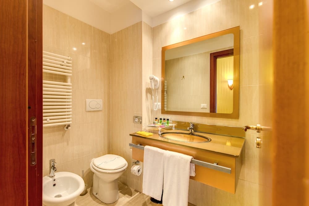 A cozy hotel bathroom features warm beige tiles, a sleek sink with a large mirror, a bidet, a toilet with a towel warmer, and neatly arranged toiletries, ideal for travelers seeking comfort and convenience.