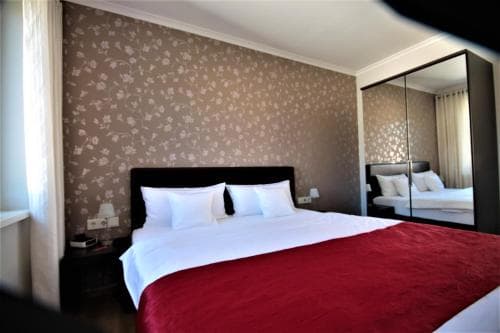 A cozy hotel room features a neatly made bed with white linens and a red blanket, set against floral wallpaper, with mirrored wardrobes reflecting the serene ambiance, ideal for travelers seeking comfort and relaxation.