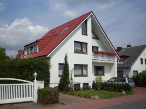 A charming white house with a red-tiled roof stands amidst lush greenery in a serene suburban neighborhood, featuring a welcoming white gate and blooming flowers, ideal for a peaceful family vacation.