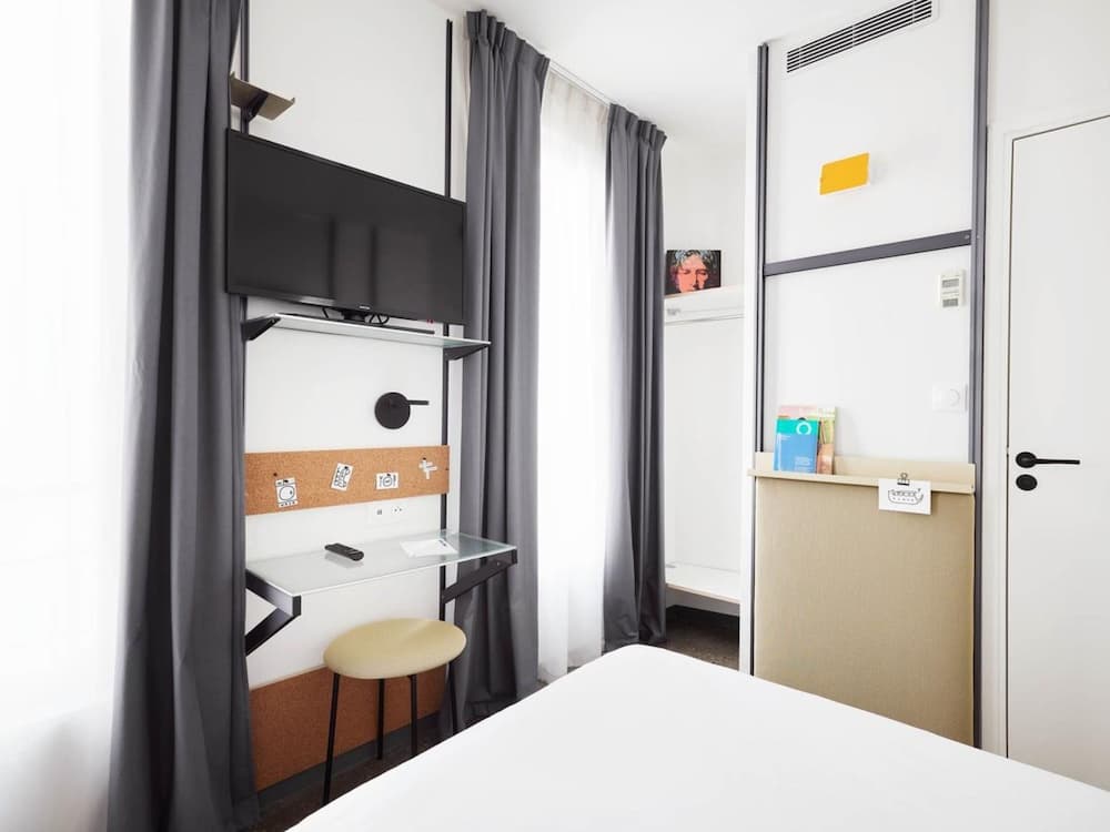 A cozy hotel room features a minimalist desk setup with a mounted TV, soft lighting through sheer curtains, and personal touches like a corkboard and small shelf, ideal for travelers seeking comfort and functionality in a compact space.