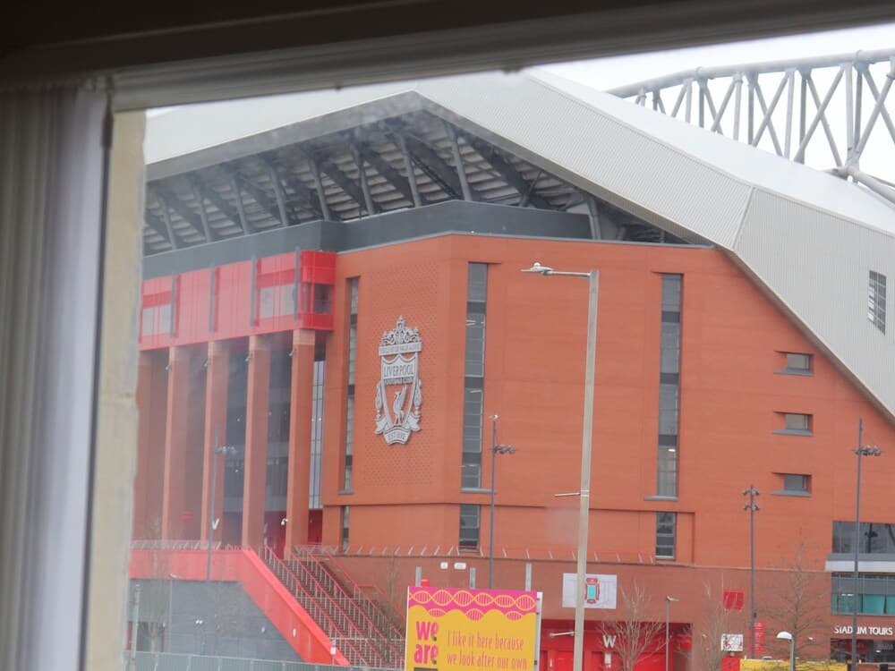 A vibrant view of a football stadium's exterior through a window, featuring bold red architecture, a prominent club crest, and signage for stadium tours, evokes excitement for sports enthusiasts planning their visit.