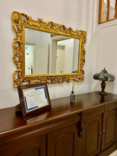 An elegant hallway features a grand golden-framed mirror above a polished wooden cabinet adorned with a certificate and decorative lamp, evoking a classic and sophisticated atmosphere ideal for boutique hotel or heritage site exploration.