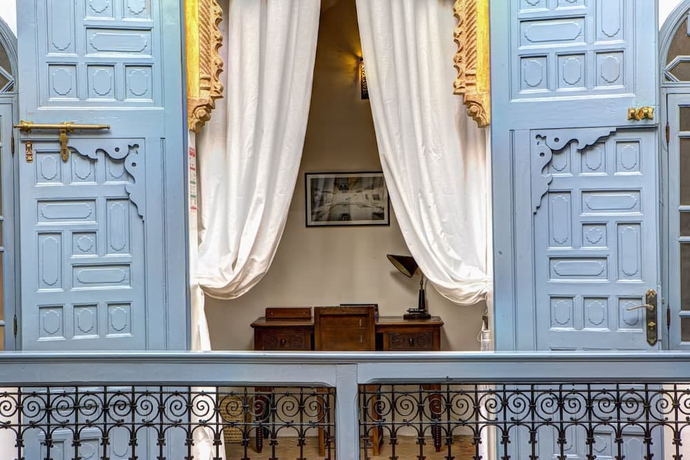 A charming balcony view of an ornate interior framed by blue doors and white curtains, featuring vintage furniture and a decorative railing, evokes a nostalgic atmosphere ideal for heritage tourism exploration.
