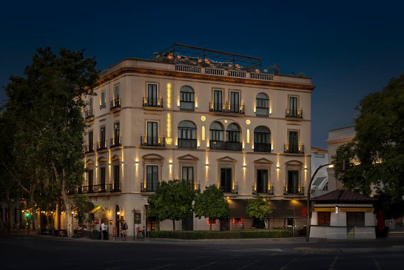 A warmly lit hotel facade in Sevilla's historic Casco Antiguo glows invitingly at dusk, featuring elegant balconies and arched windows, ideal for travelers seeking a luxurious stay in the heart of Spain's vibrant culture.