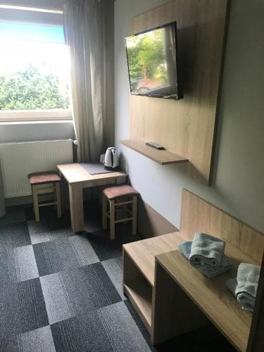 A cozy hotel room features a small wooden desk with stools by a window overlooking greenery, a mounted TV, and a bench with towels, ideal for travelers seeking comfort and functionality.