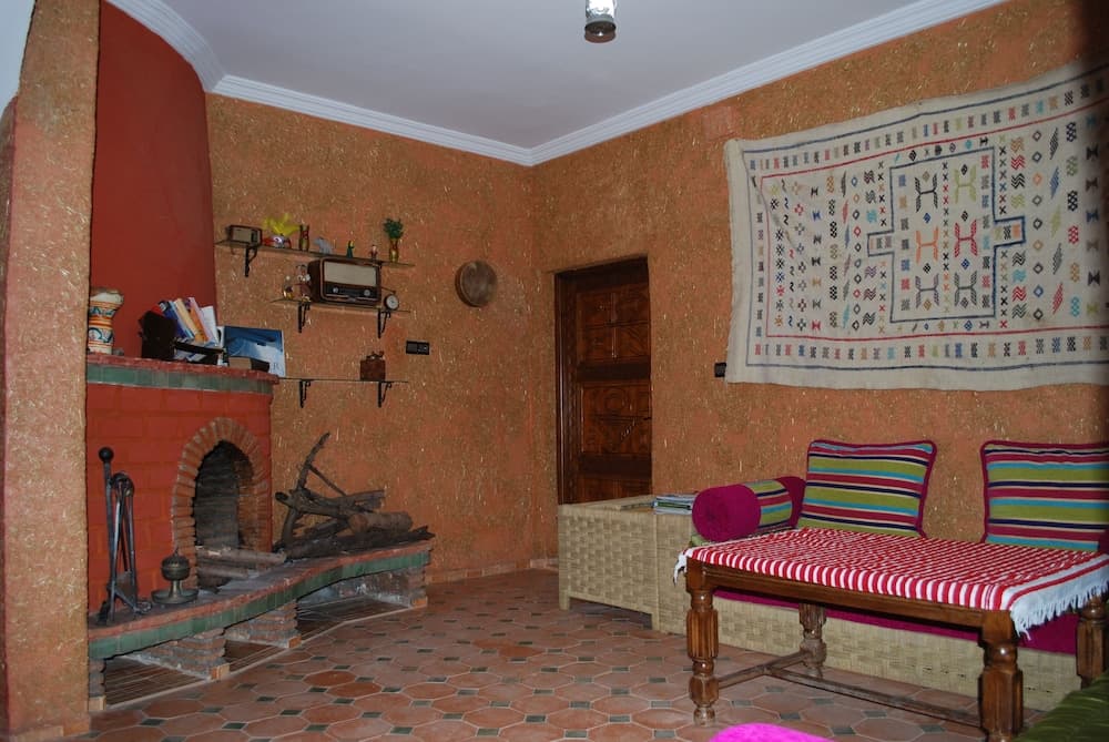 A cozy indoor lounge features terracotta walls, a tiled fireplace with logs, a woven bench adorned with colorful cushions, and a vibrant tapestry, ideal for travelers seeking a warm, rustic retreat.
