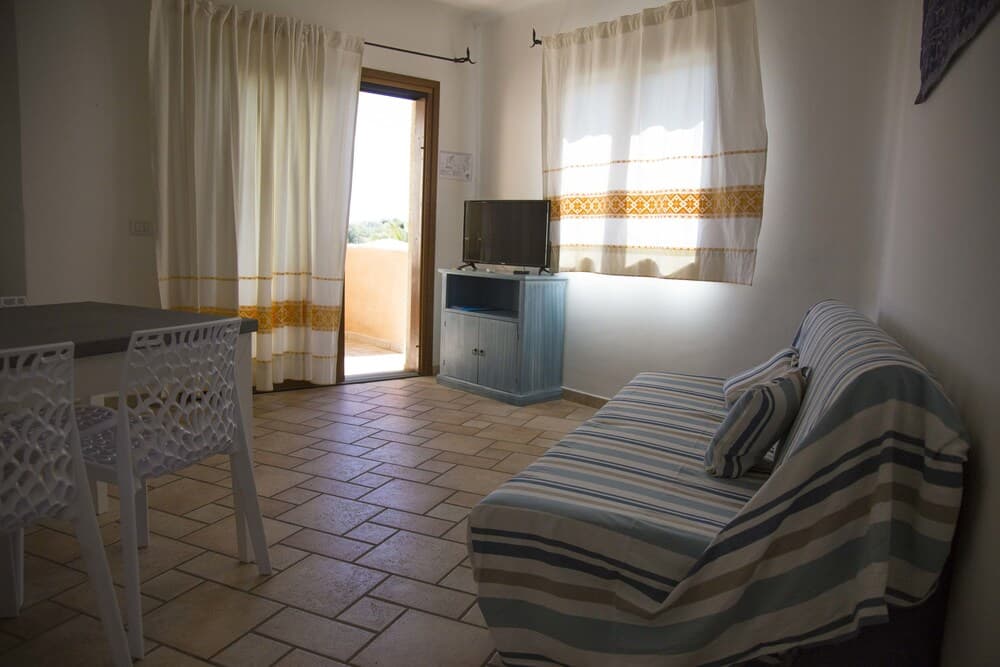 A cozy vacation rental living room features tiled floors, striped furniture, a small dining area, and bright natural light streaming through curtains, ideal for travelers seeking comfort and simplicity.