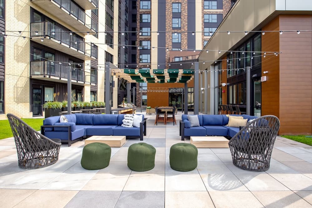 A cozy urban courtyard features modern blue sofas, green poufs, and wicker chairs under string lights, creating an inviting atmosphere ideal for city dwellers seeking relaxation or social gatherings.